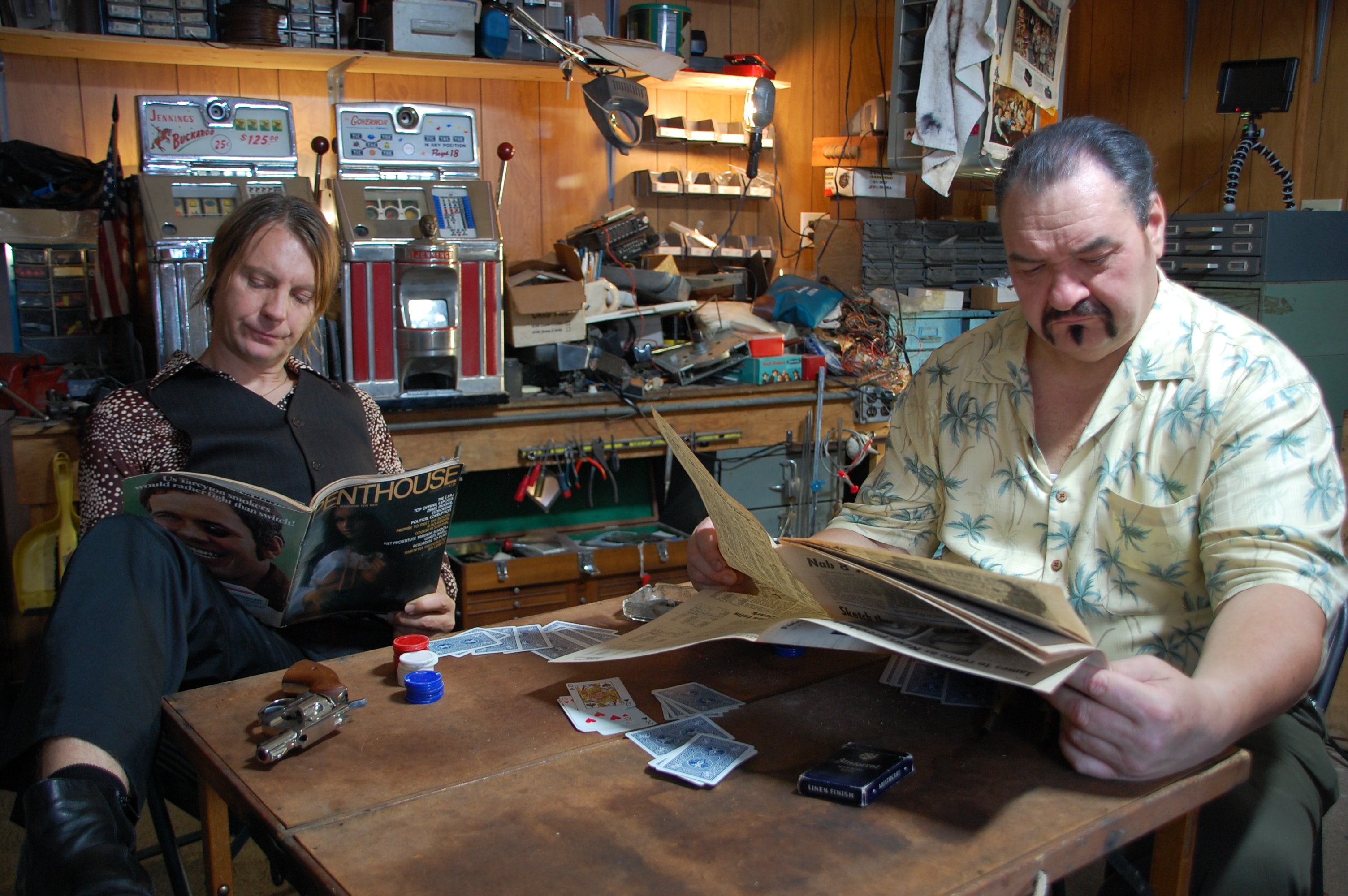 Two men reading magazines at a table covered in playing cards in a workshop