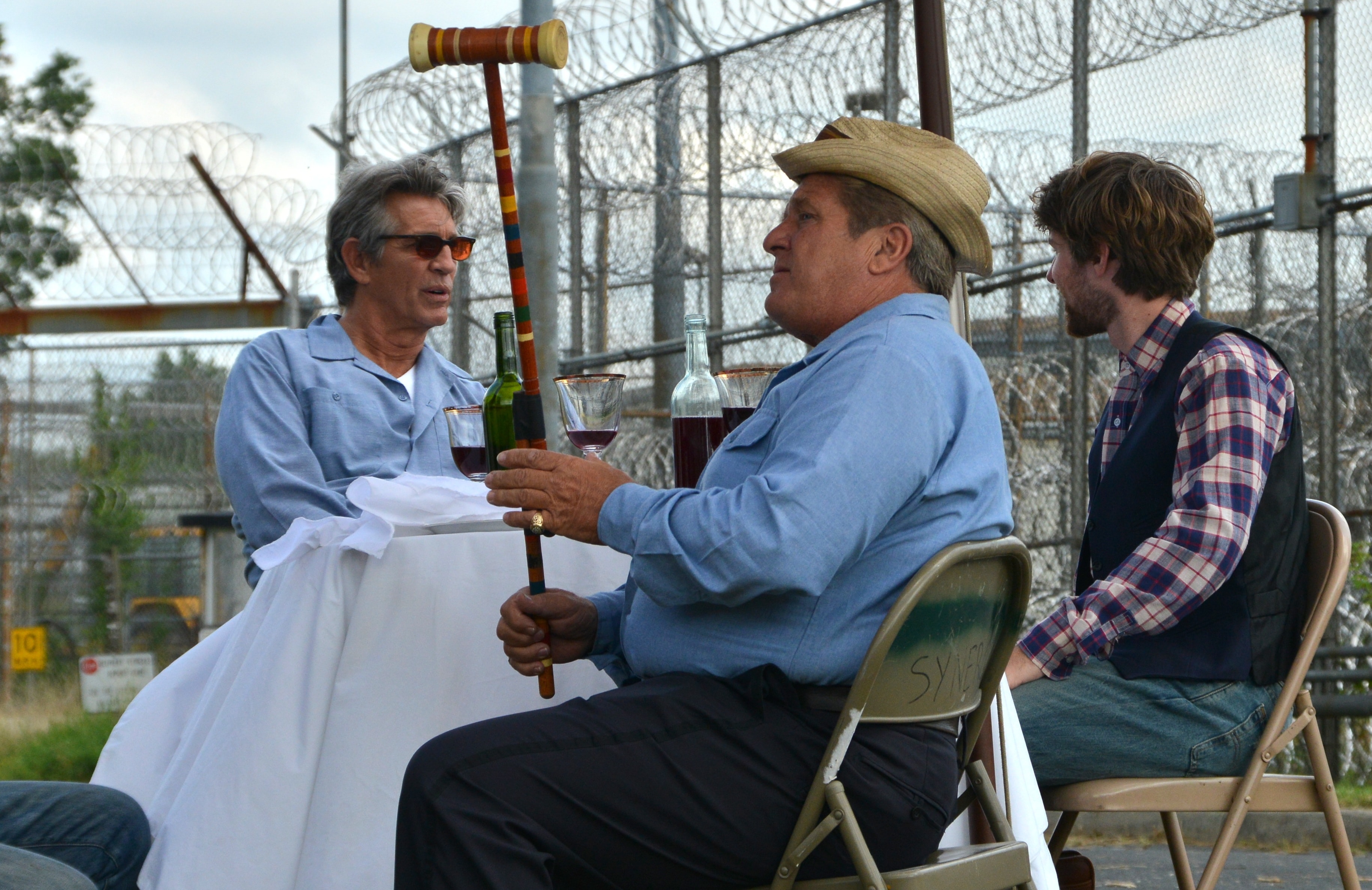 Three men drinking wine at a table outside a prison fence