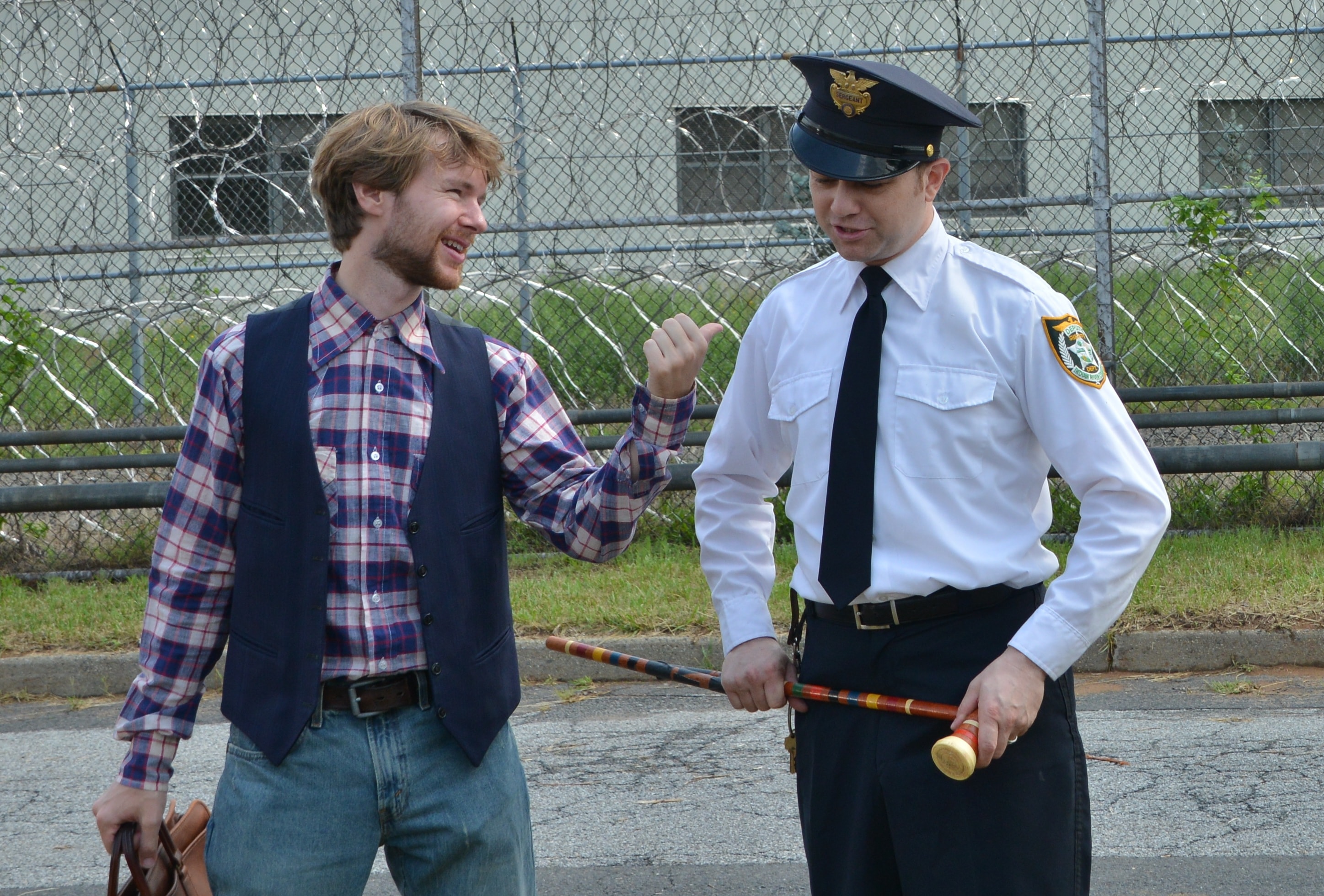 A young man in a plaid shirt laughs with a guard holding a cane outside a prison