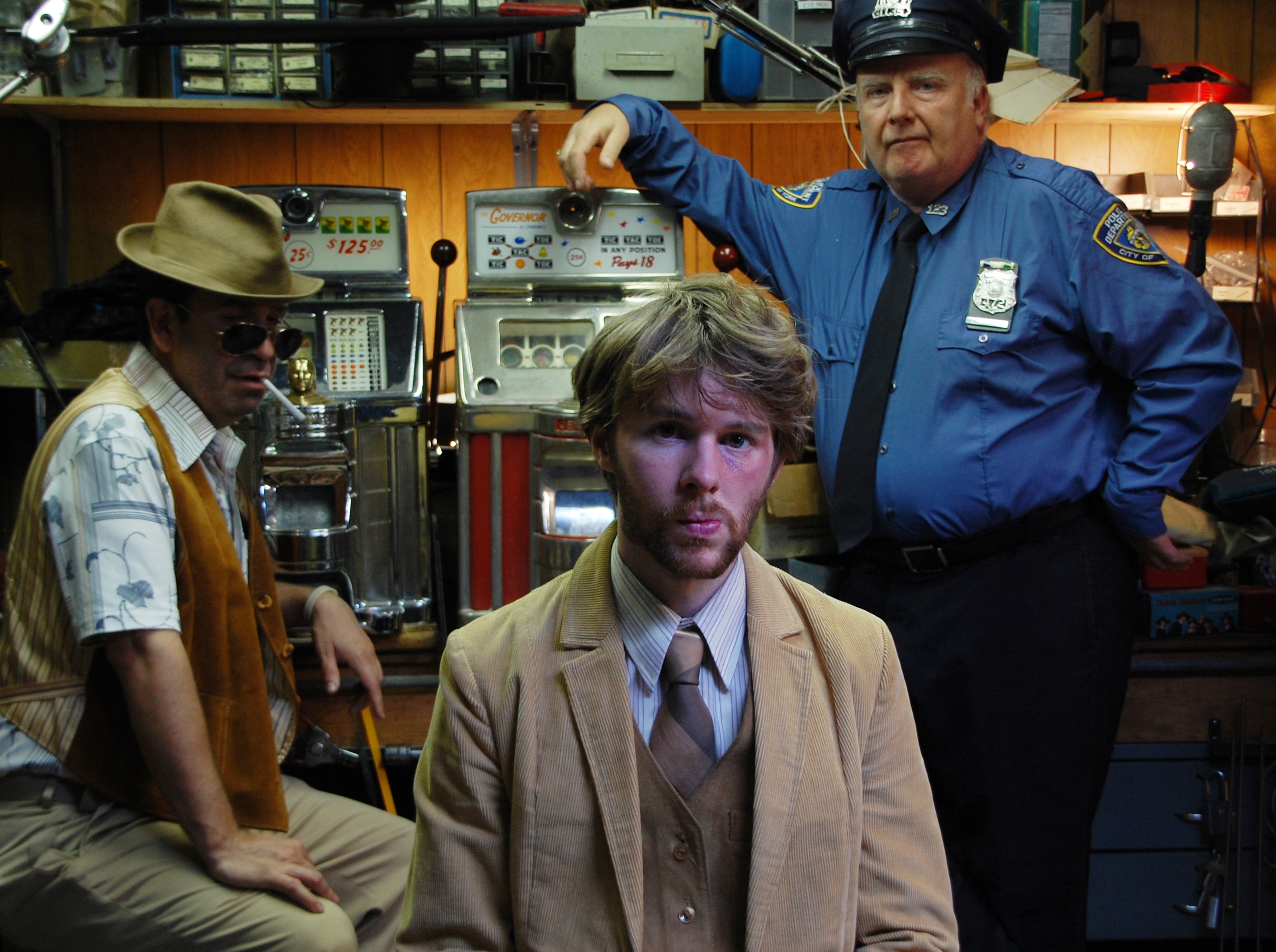 A young man in a tan suit sits between a man in a hat and a police officer