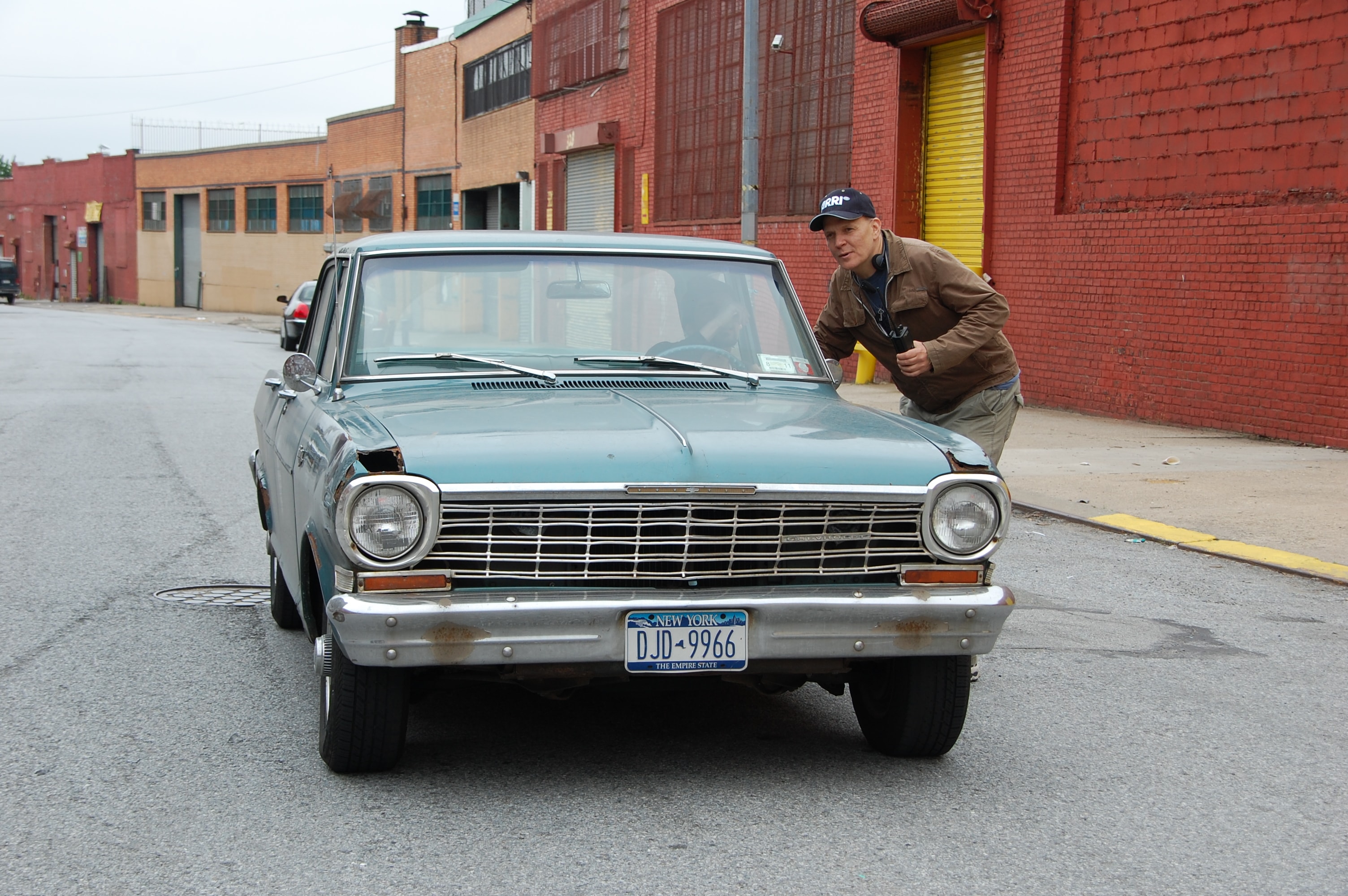 A man in a cap poses behind a vintage teal Chevy Nova on a Brooklyn street