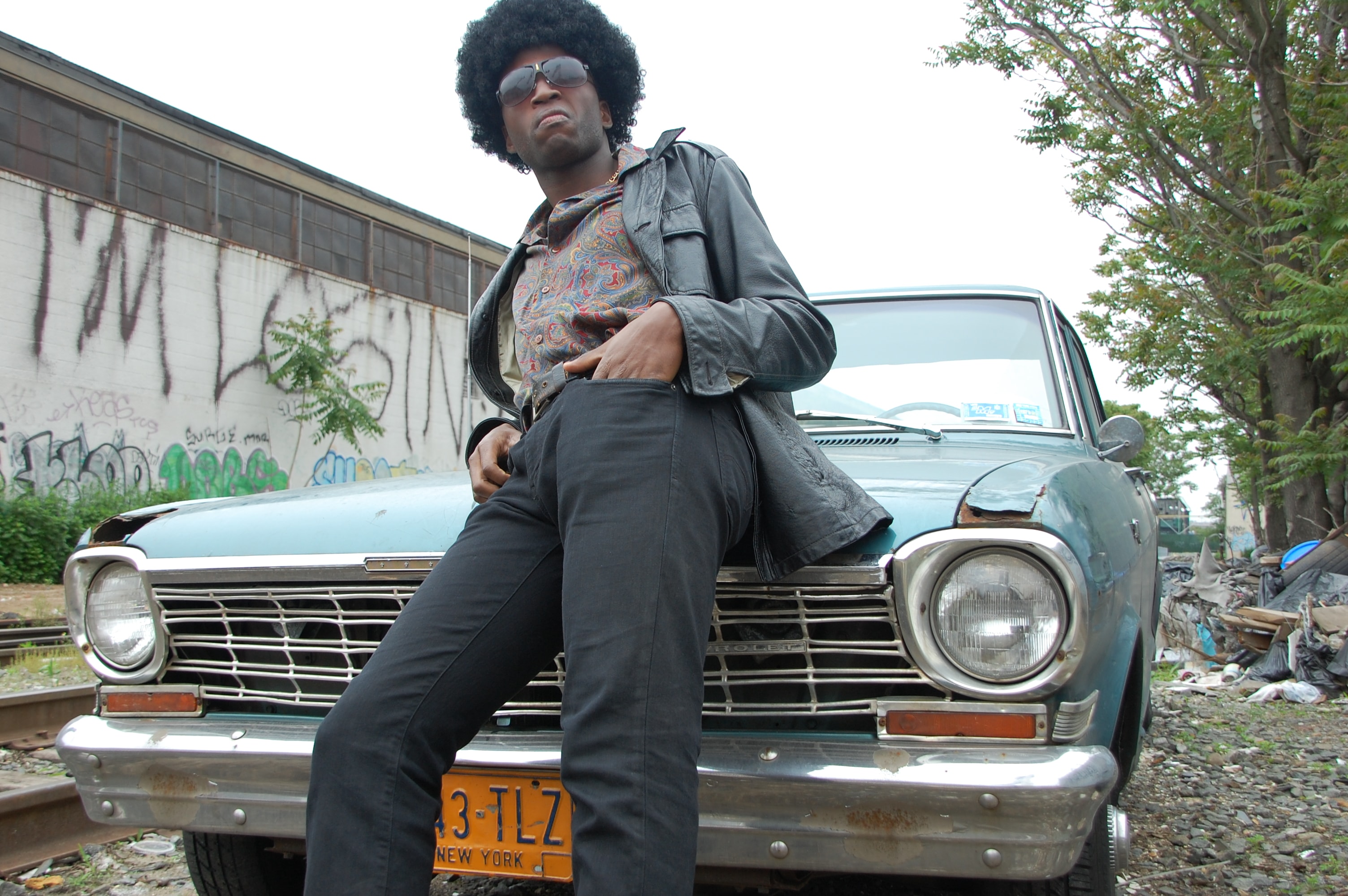 A man in an afro and leather jacket leans against a vintage car in front of graffiti