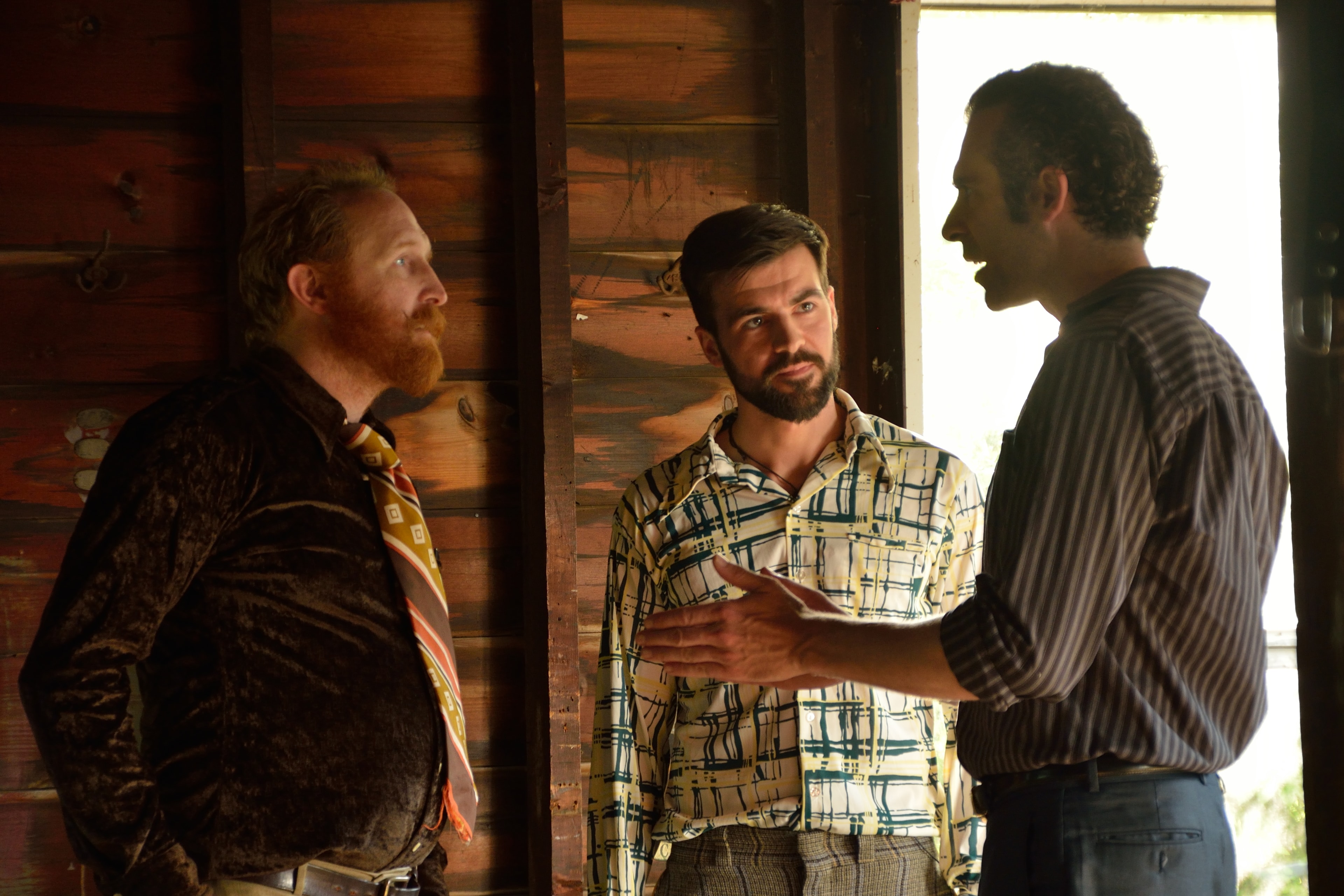 Three men in 1970s clothing in conversation inside a wooden doorway
