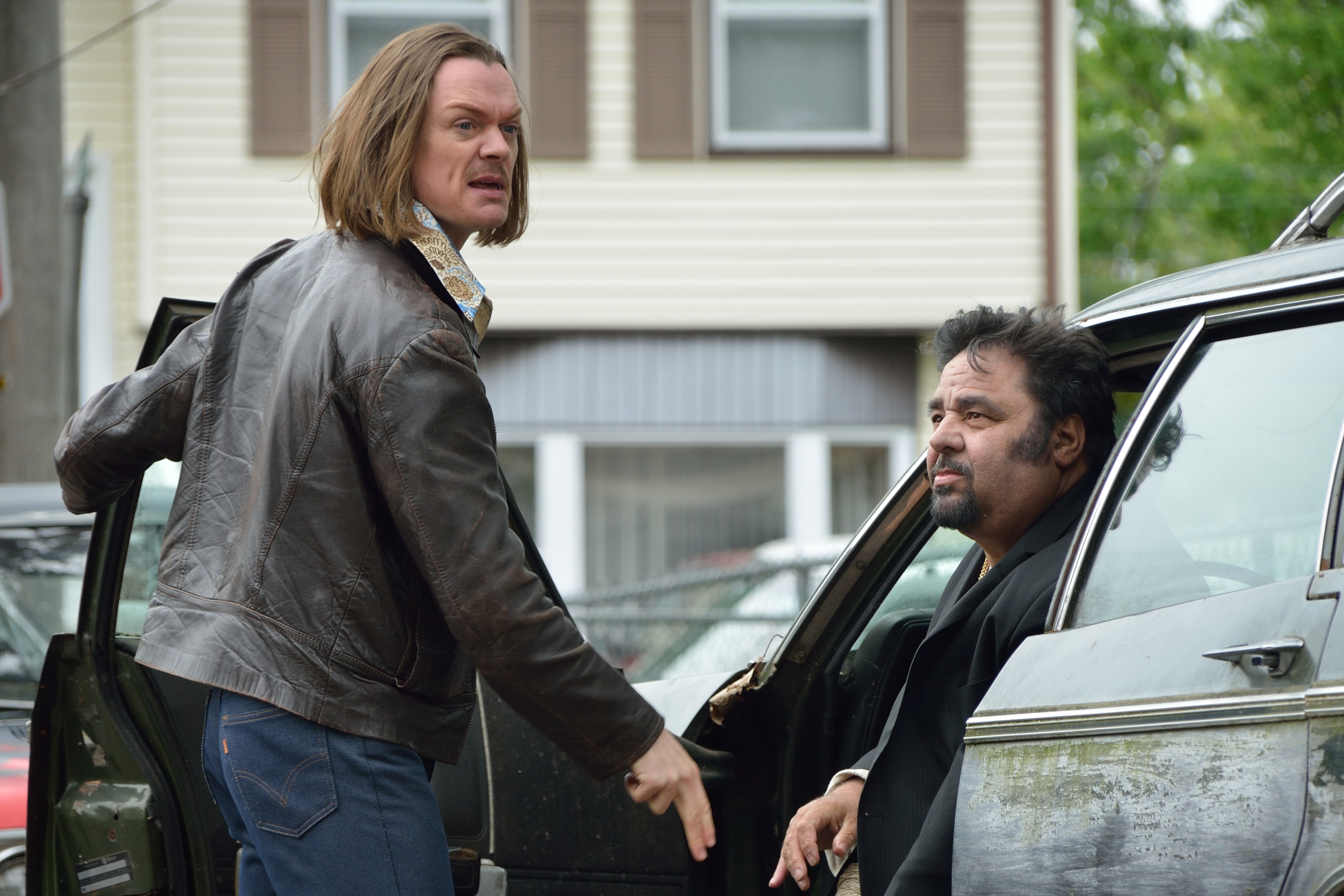 A man with long hair confronts another man getting out of a car on a suburban street