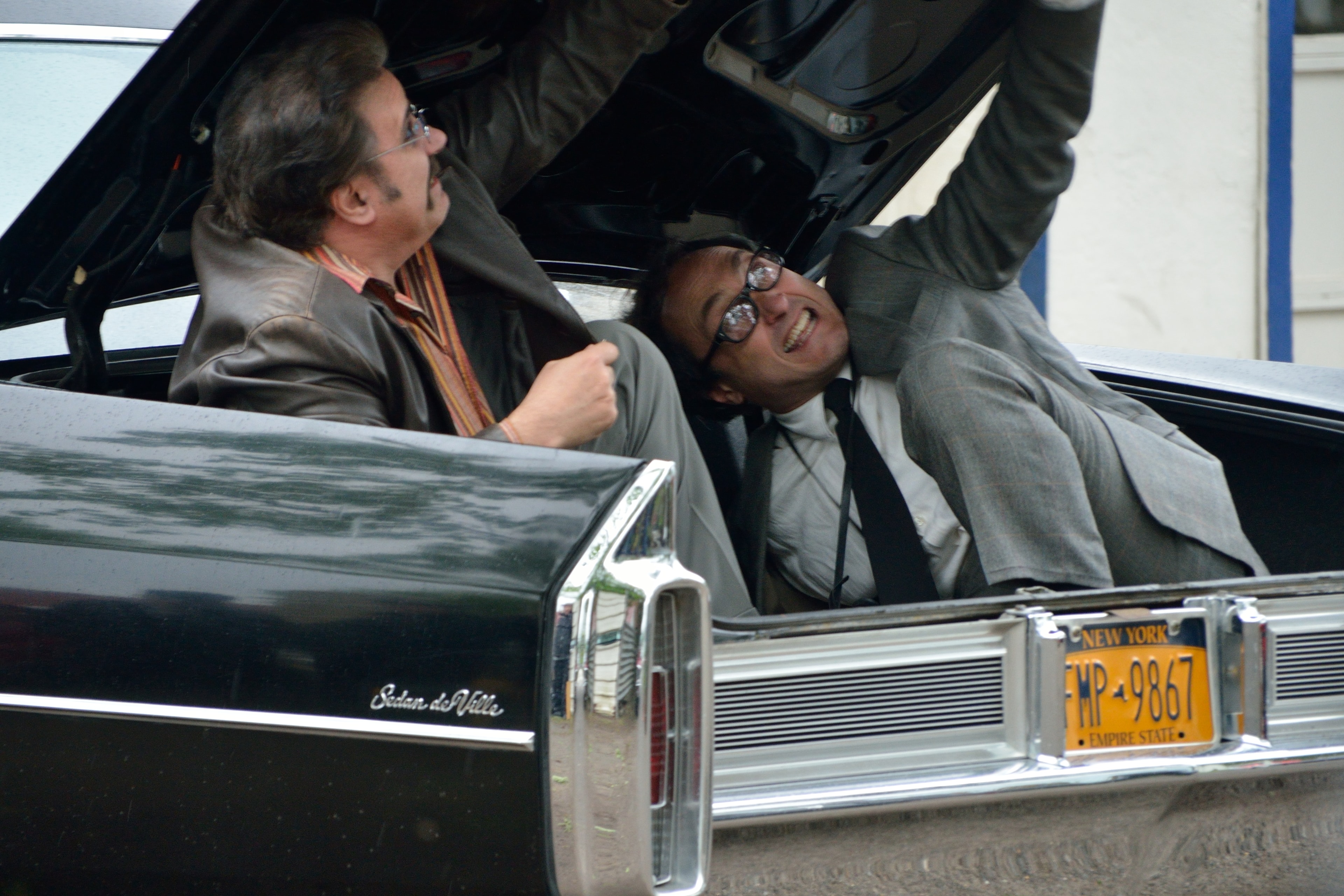 Two men laugh while one is stuffed into the trunk of a Cadillac Sedan de Ville