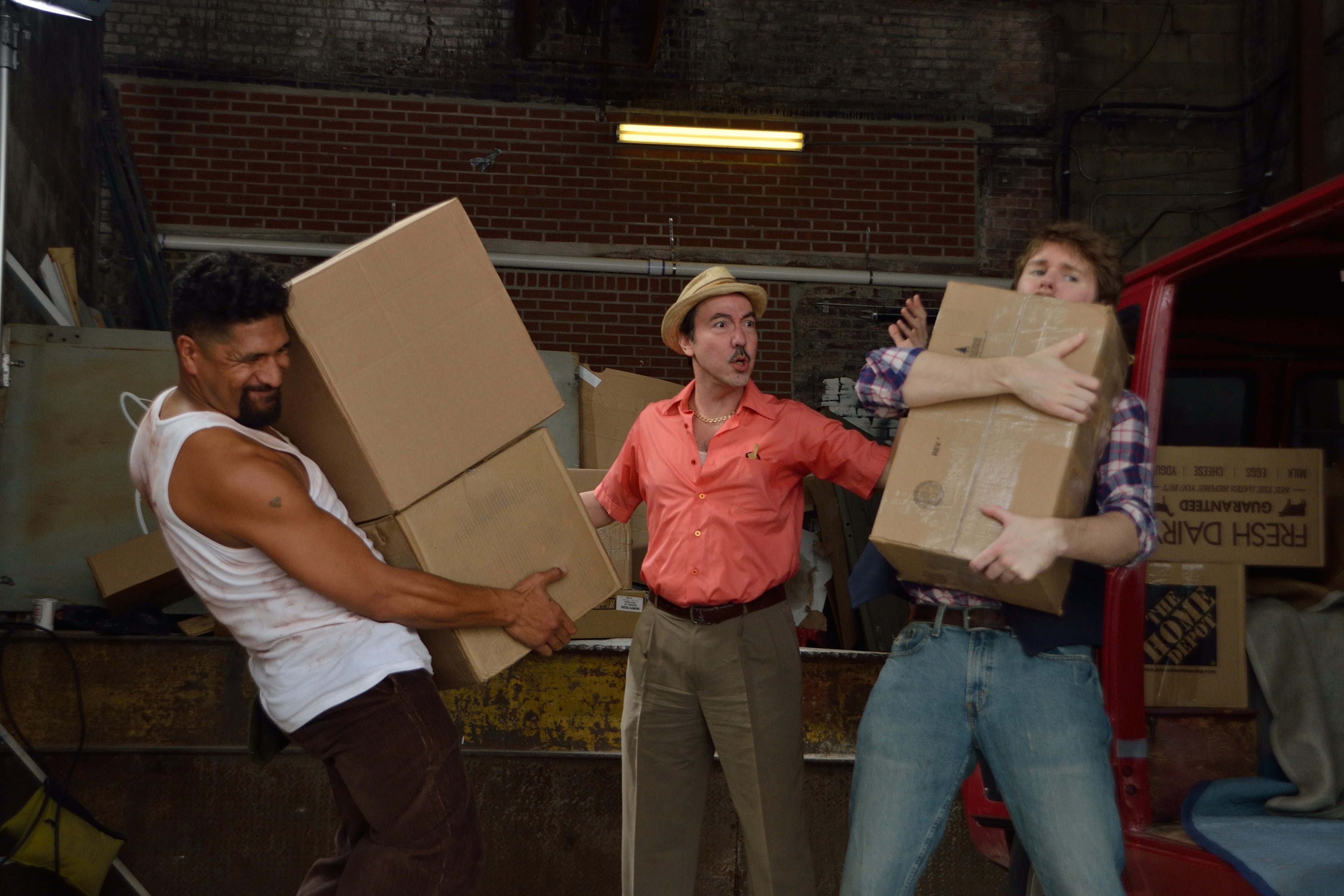 Three men in 1970s costumes carry cardboard boxes in a warehouse