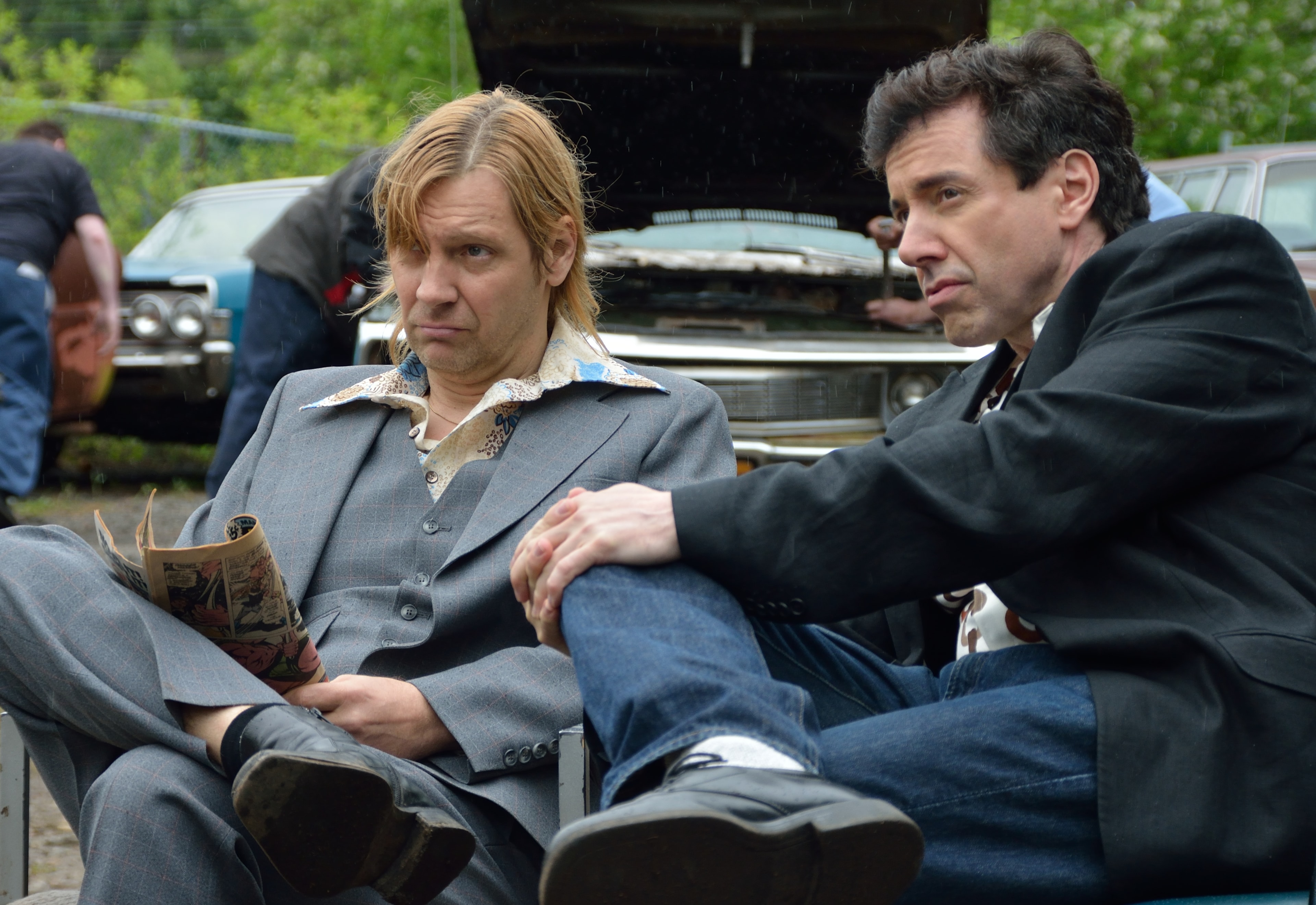 Two men in 1970s suits sit together in a junkyard with vintage cars behind them