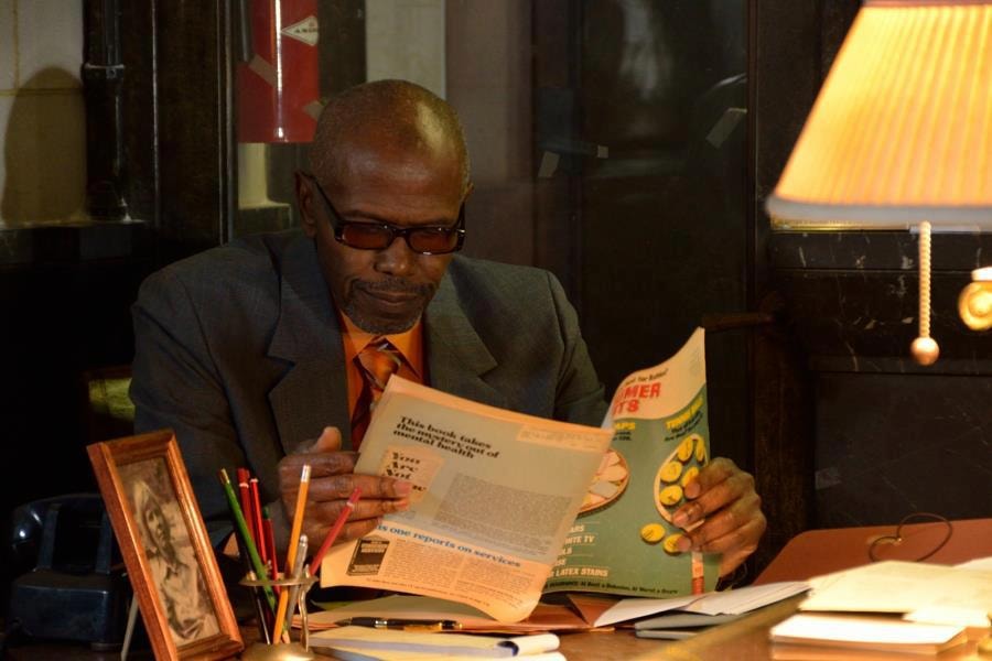 A man in a grey suit and tinted glasses reads a magazine at a warmly lit desk