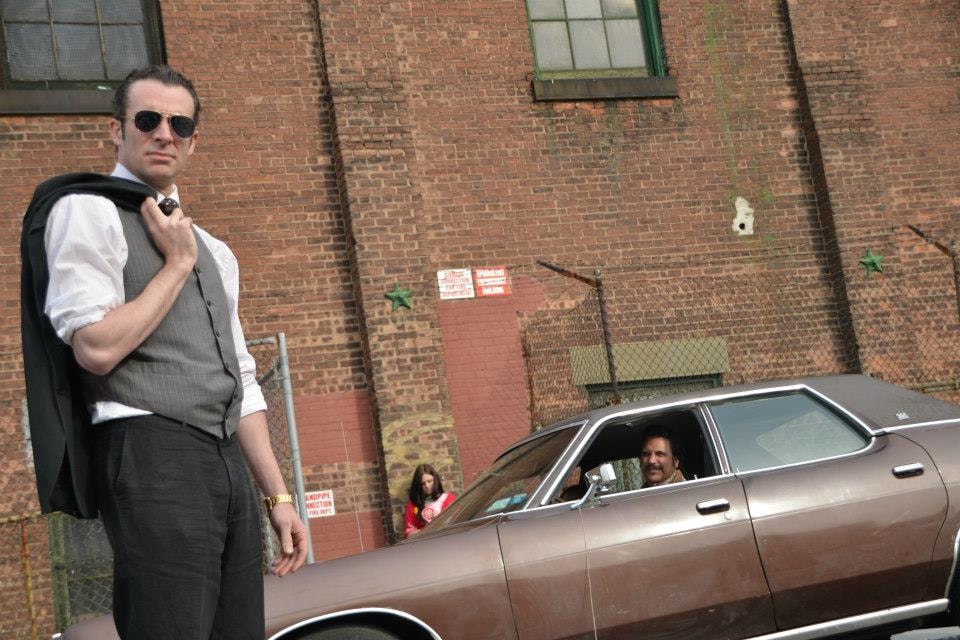 A man in a vest and sunglasses stands in front of a brick wall with a vintage car