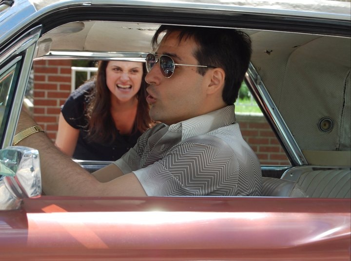A woman leans into a vintage car window arguing with a man in aviator sunglasses