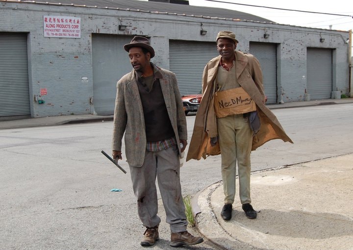 Two men walk a Brooklyn street, one holding a cardboard sign reading Need Money