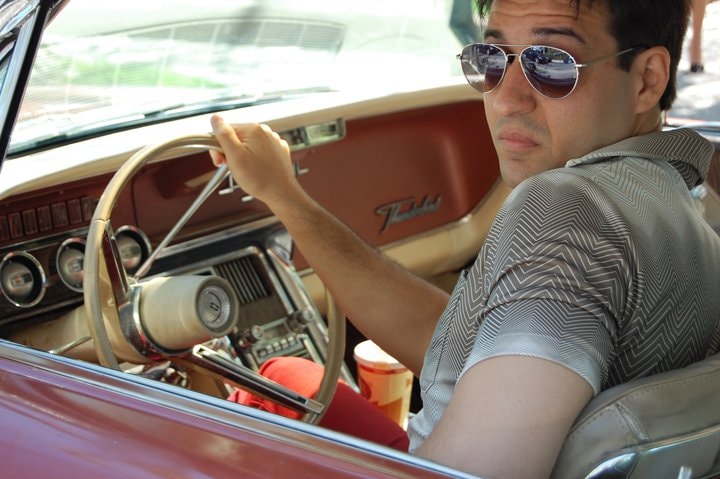 A man in aviator sunglasses grips the wheel of a vintage Ford Thunderbird