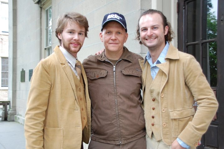 Three men in 1970s clothing stand outside a building on a city street