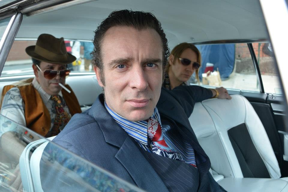 A man in a blue suit stares directly at camera from inside a car